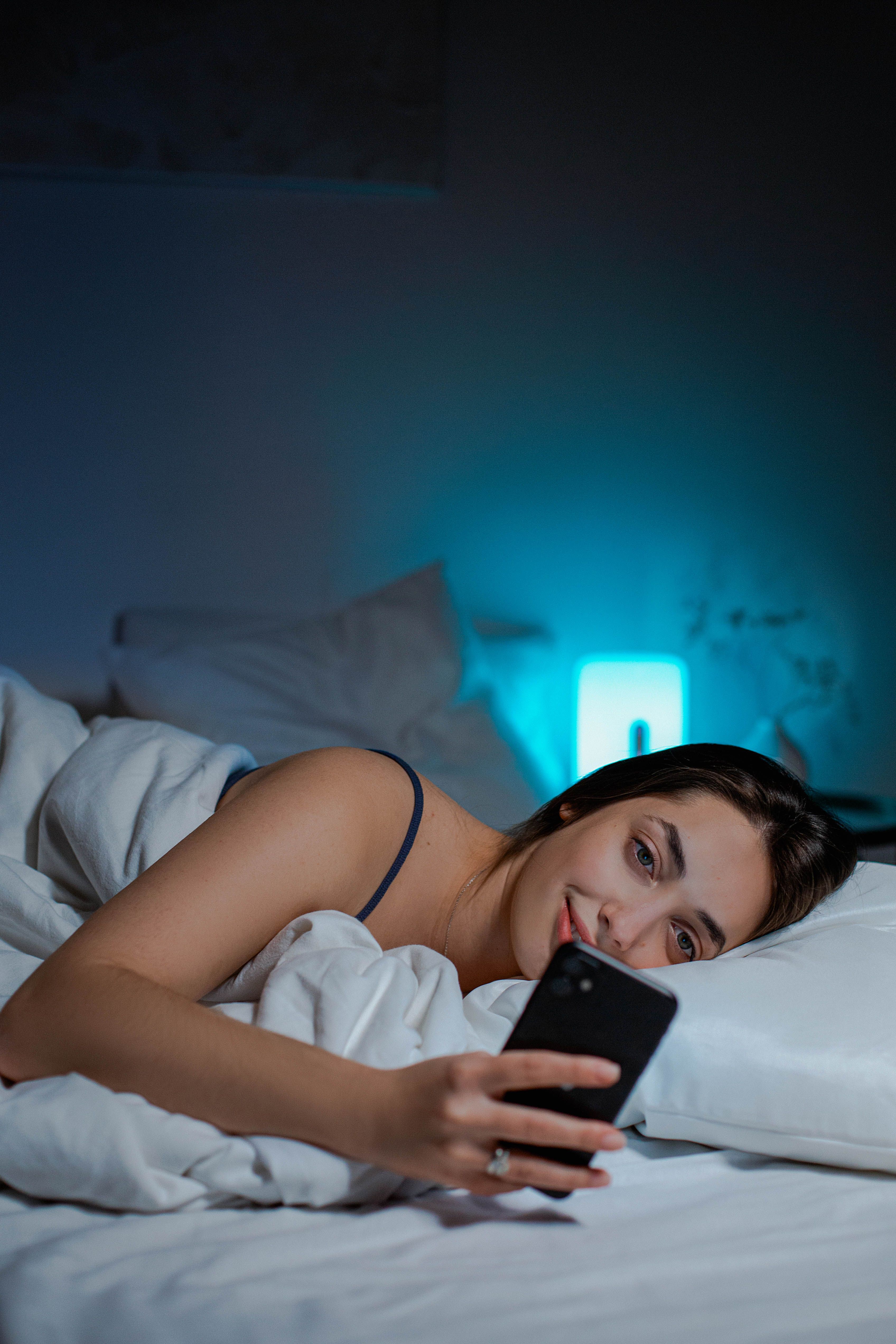 A woman watching her phone while in bed with a lamp switched on behind her | Source: Pexels