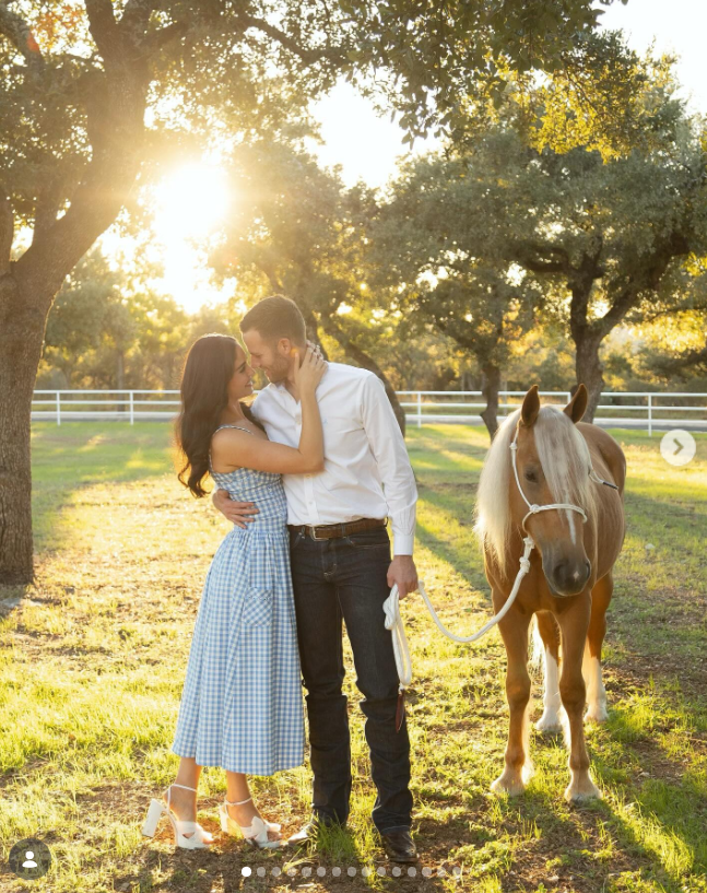 Juliette Dell embraces her husband, Chase Chamberlin, in a sunlit pasture during golden hour as he holds the lead rope of a palomino pony standing beside them. Juliette wears a blue gingham midi dress and white platform heels, smiling up at Chase as he leans in close in a crisp white button-down shirt, dark jeans, and boots. | Source: Instagram/juliette_dell