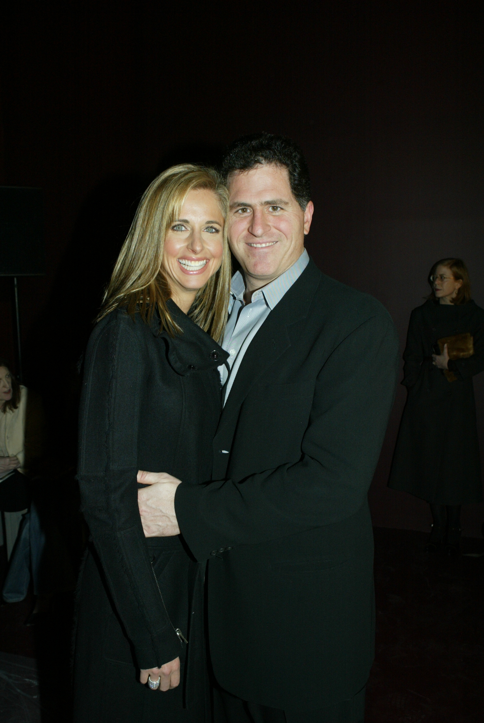 Susan Dell and Michael Dell smiled warmly while embracing at the Fall 2004 Phi fashion show in New York on February 13, 2004. Susan wore a stylish long black coat with a wide collar, while Michael coordinated beside her in a classic black blazer over an open-collar grey dress shirt. | Source: Getty Images