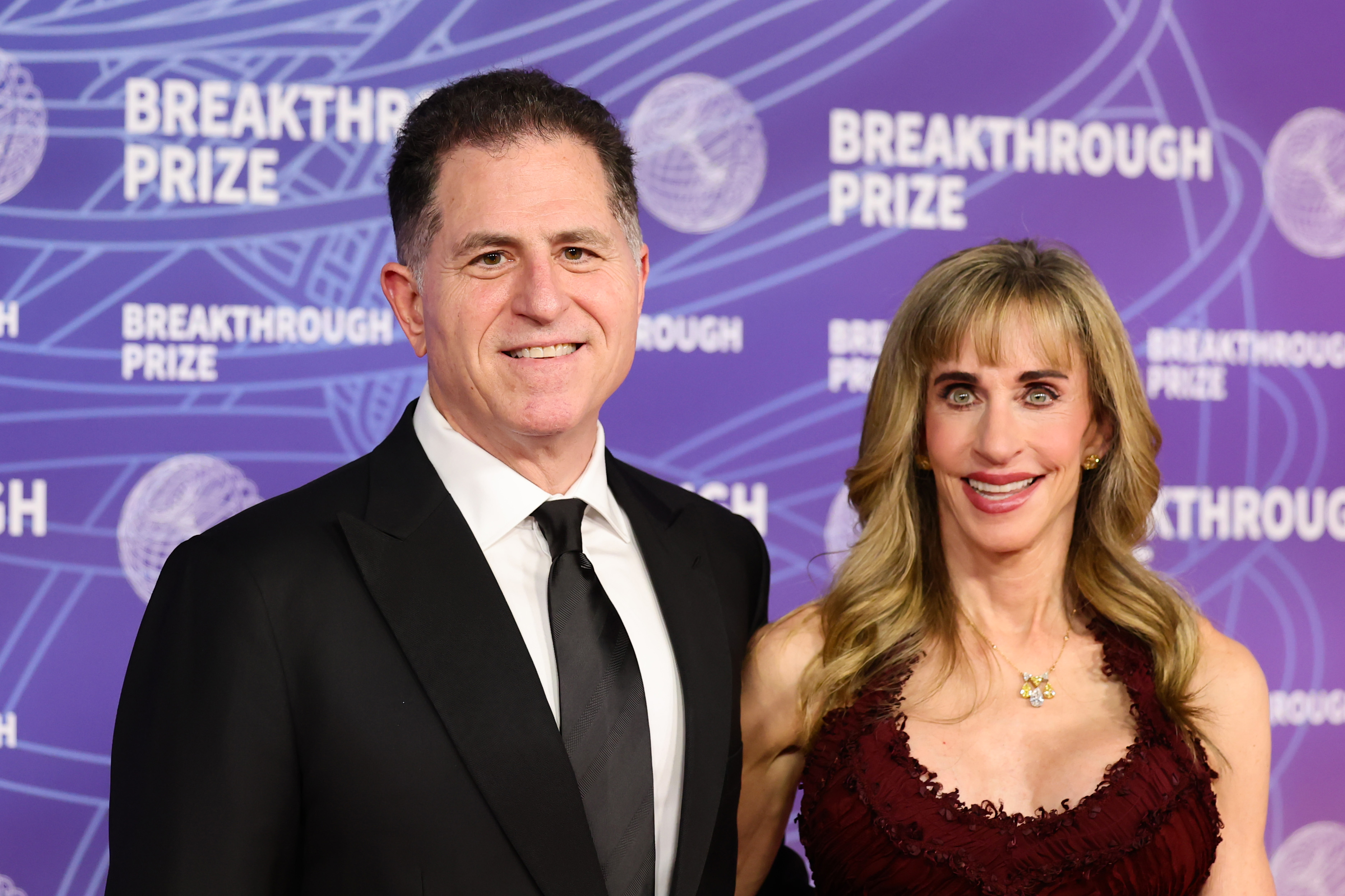 Michael Dell and Susan Dell smile together on the red carpet at the 12th Breakthrough Prize Ceremony at Barker Hangar on April 18, 2026, in Santa Monica, California. Michael dressed in a classic black tuxedo, white shirt, and black necktie, while Susan wore a sleeveless deep-burgundy gown featuring ruffled texture, accessorized with button earrings and a delicate pendant necklace. | Source: Getty Images