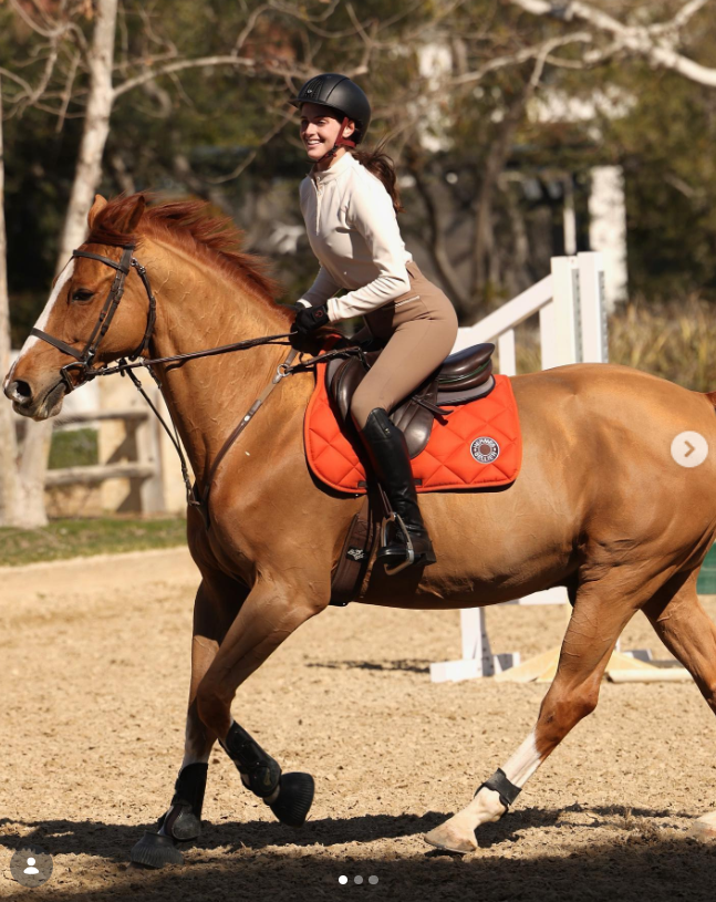 Alexa Dell smiles as she rides a chestnut horse in an equestrian arena, dressed in riding gear that includes a helmet, cream zip-up top, tan breeches, and tall black riding boots. | Source: Instagram/alexakdell