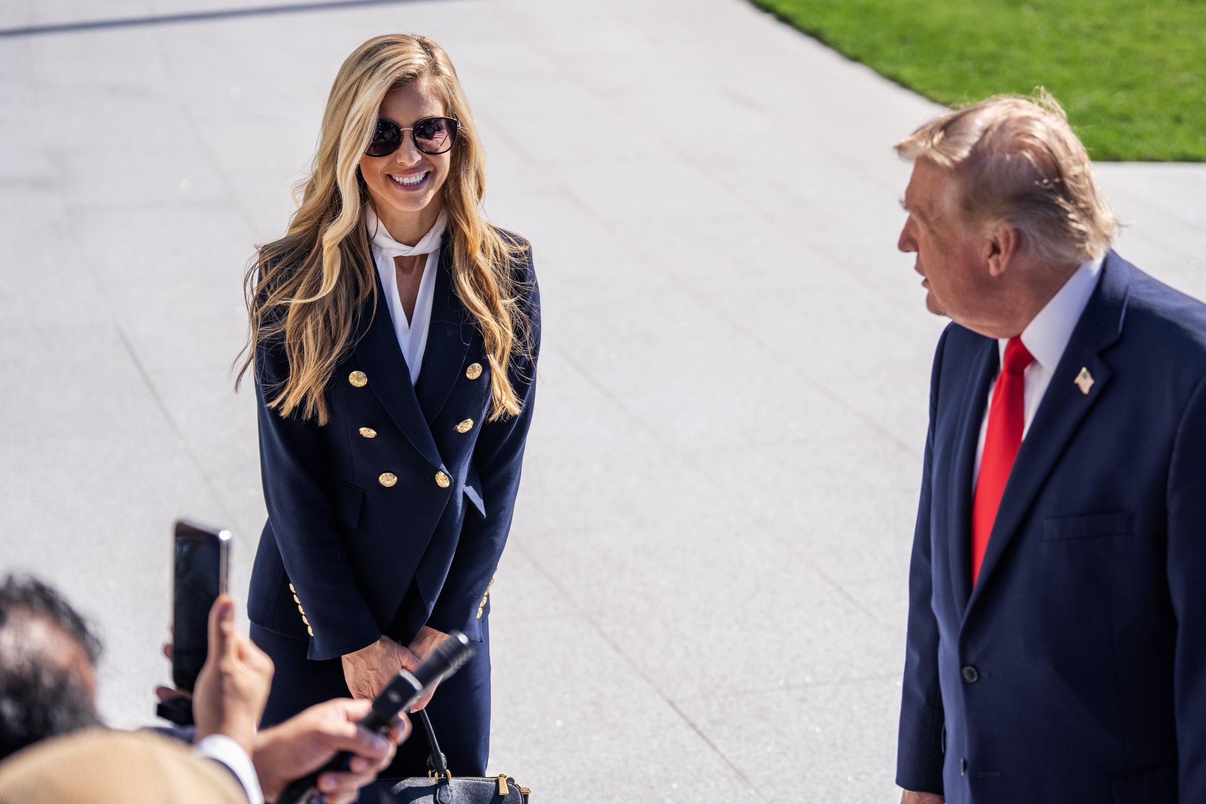 White House Press Secretary Katie Zacharia and President Donald Trump speak with reporters on the South Lawn of the White House before departing aboard Marine One on October 13, 2026, in Washington, D.C. Zacharia smiles in her navy double-breasted suit with gold buttons, dark sunglasses, and a black handbag, standing attentively beside Trump as members of the press gather microphones and phones to capture their remarks. | Source: Getty Images