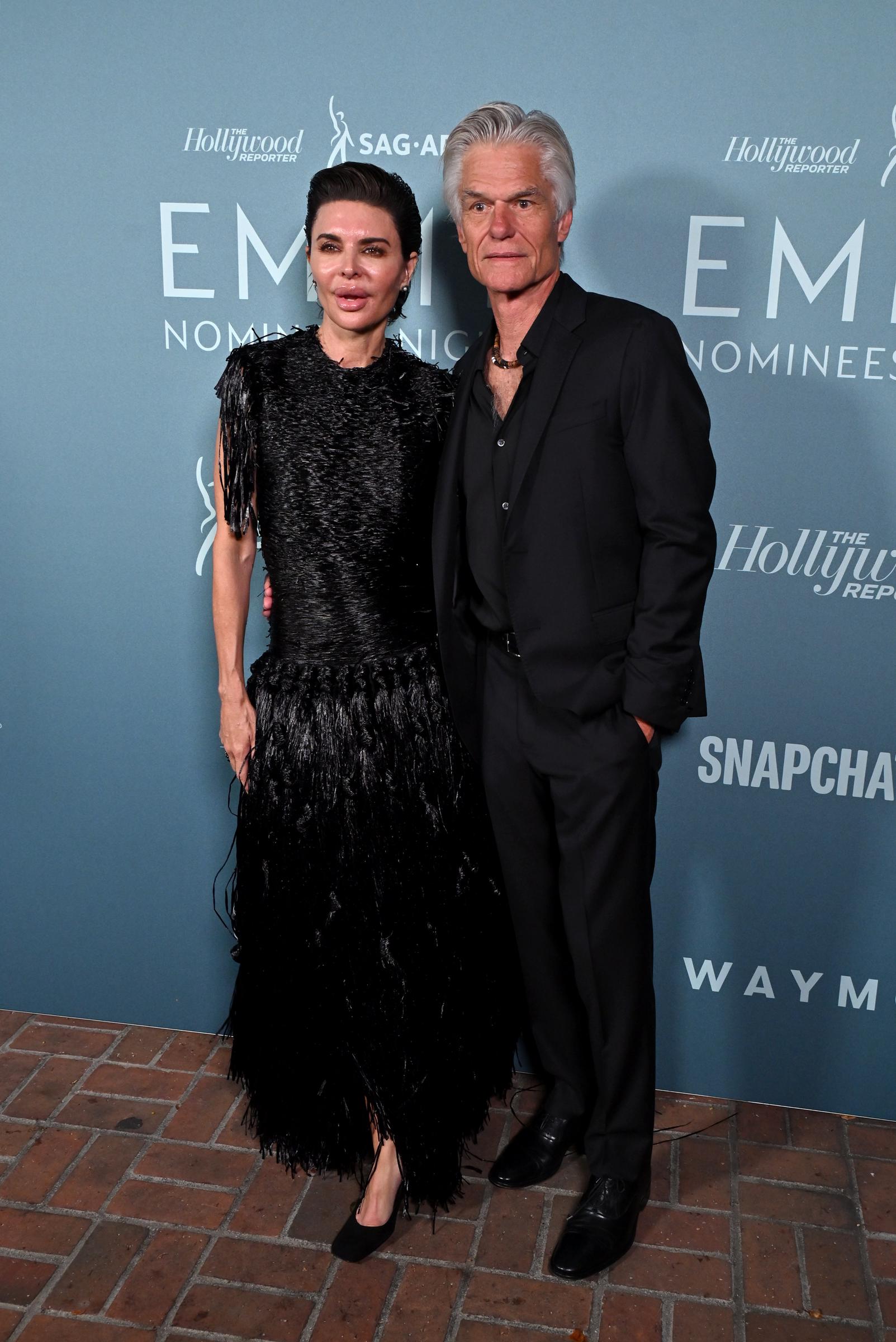 Lisa Rinna and Harry Hamlin pose together in front of the Emmy Nominees Night backdrop, with Rinna showcasing her dramatic black fringe gown and Hamlin complementing her in a sleek all-black suit. | Source: Getty Images