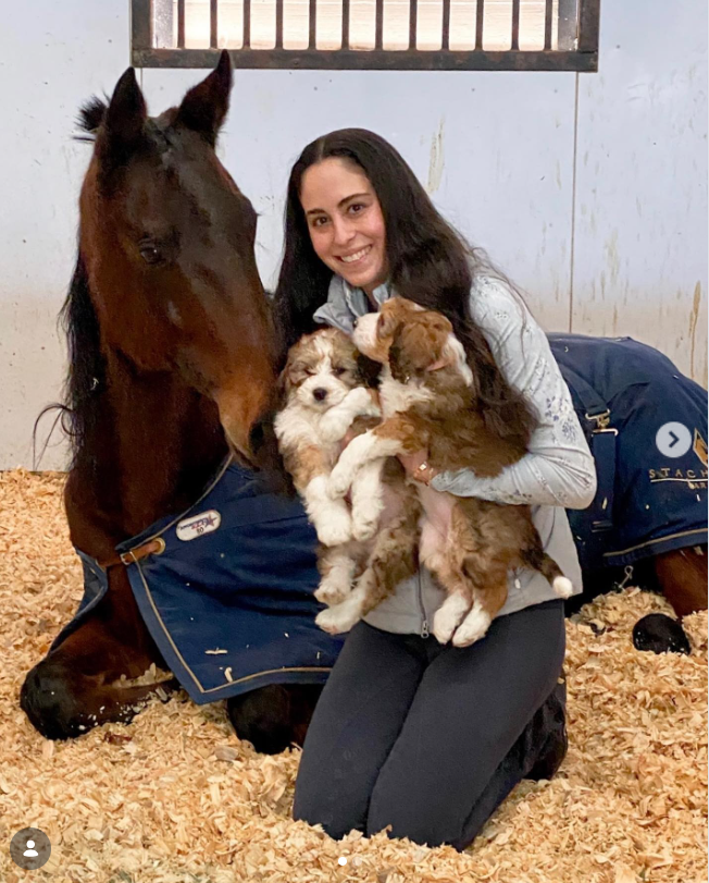 Juliette Dell kneels in a stable stall while happily cuddling two fluffy puppies in her arms, accompanied by a blanketed horse resting comfortably behind her in the wood shavings. | Source: Instagram/juliette_dell