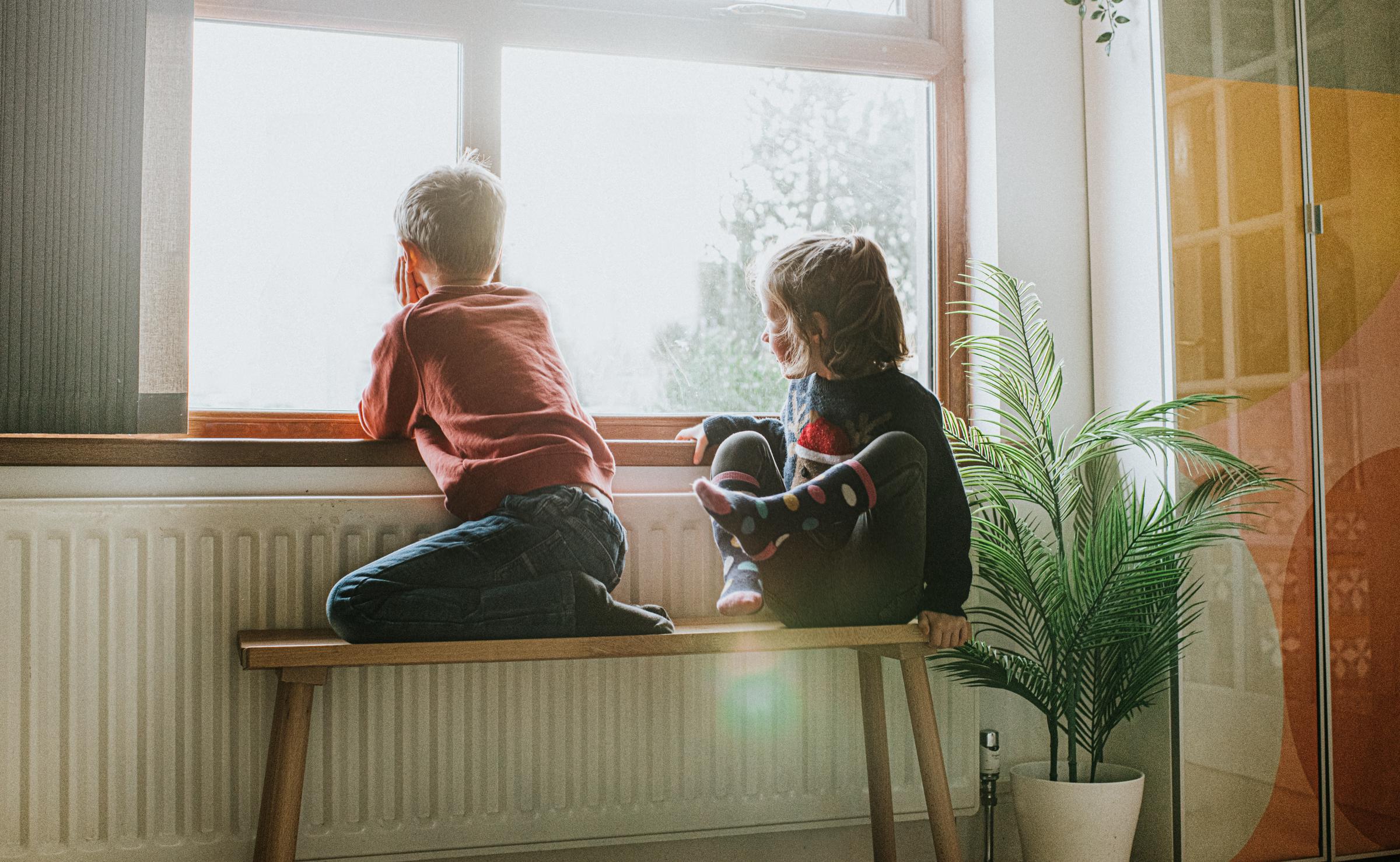 A young girl and boy sit on a wooden bench beside a long radiator and a sunny window | Source: Getty Images