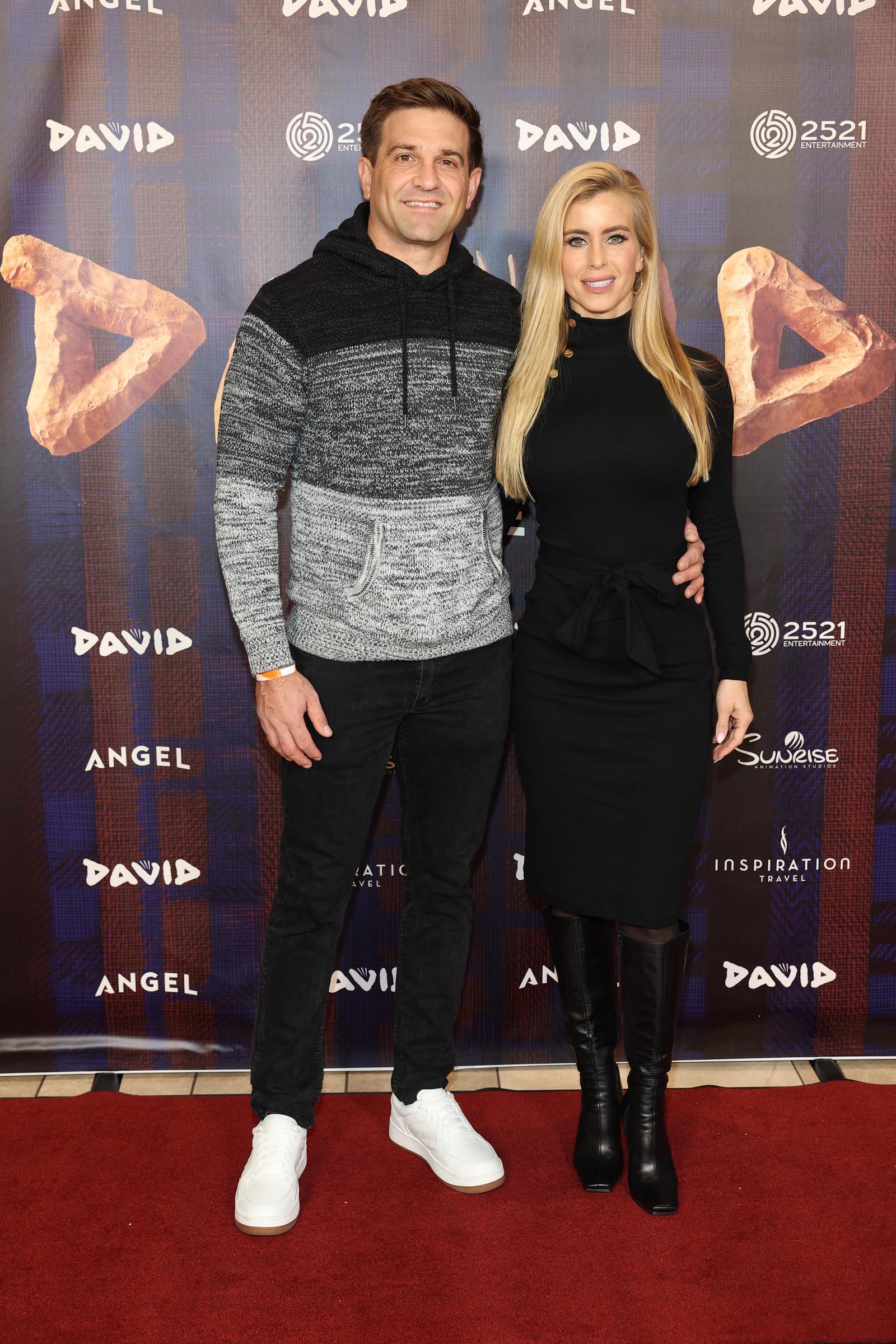 Tyler Zacharia and Katie Zacharia pose full-length on the red carpet at the Nashville premiere of "David" at AMC DINE-IN Thoroughbred 20 on December 14, 2025, in Franklin, Tennessee. Showcasing their footwear and full silhouettes against the Angel Studios step-and-repeat backdrop, Tyler pairs clean white low-top sneakers with his two-toned hooded knit sweater and black denim, while Katie finishes her belted knee-length black dress with polished black knee-high leather boots. | Source: Getty Images