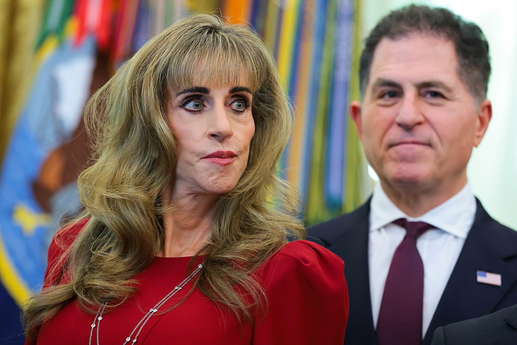 Dell Technologies CEO Michael Dell and his wife, Susan Dell, look on during an event in the Oval Office at the White House on October 7, 2026, in Washington, D.C. Susan wears a red dress with slightly gathered shoulders accented by layered silver necklaces, while Michael stands beside her in a dark suit, white collared shirt, and deep red tie with an American flag lapel pin as President Donald Trump announced the activation of automatic enrollment for the "Trump Accounts" seed deposit program. | Source: Getty Images