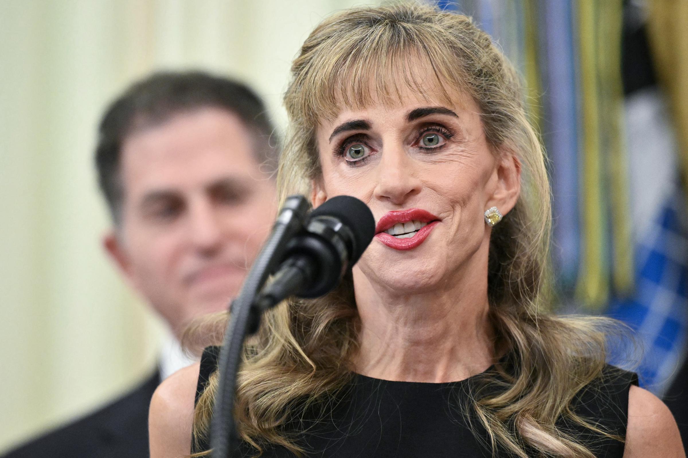 Susan Dell speaks into a microphone in the Oval Office of the White House in Washington, D.C., on July 6, 2026, during the launch of the "Trump accounts" youth investment initiative, with her husband, Dell Technologies CEO Michael Dell, looking on behind her. Susan wears a sleeveless black dress, bright red lipstick, and sparkling yellow diamond halo earrings, with her blonde-highlighted hair styled in soft waves and front bangs. | Source: Getty Images