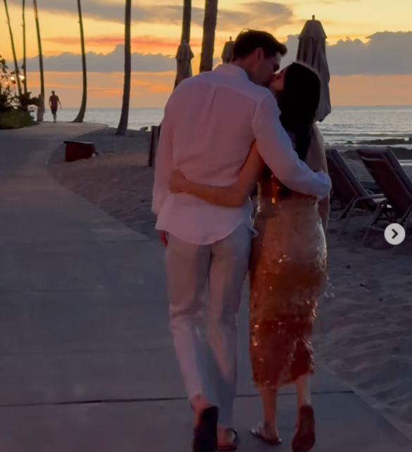 Alexa Dell and her husband, Harrison Refoua, share a kiss while walking along a seaside path at sunset, with Alexa in a sparkling sleeveless gold dress and Harrison in a white button-down shirt with light trousers. | Source: Instagram/alexakdell