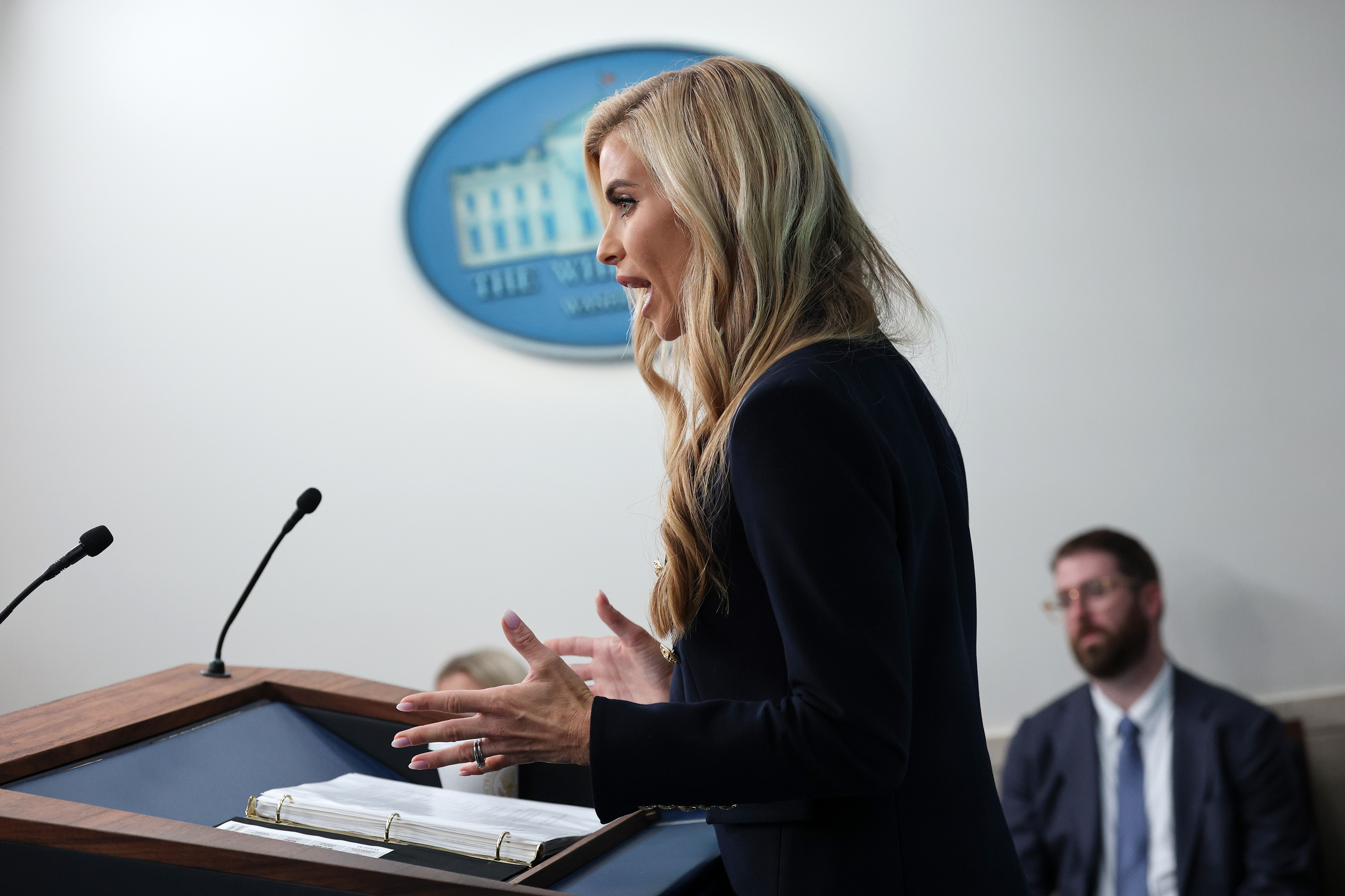White House Press Secretary Katie Zacharia speaks and gestures from the podium during her debut media briefing in the James S. Brady Press Briefing Room at the White House on October 13, 2026, in Washington, D.C. Captured in profile with notes open before her, she wears a navy-blue blazer over a white top, gesturing with both hands as reporters look on in the background. | Source: Getty Images
