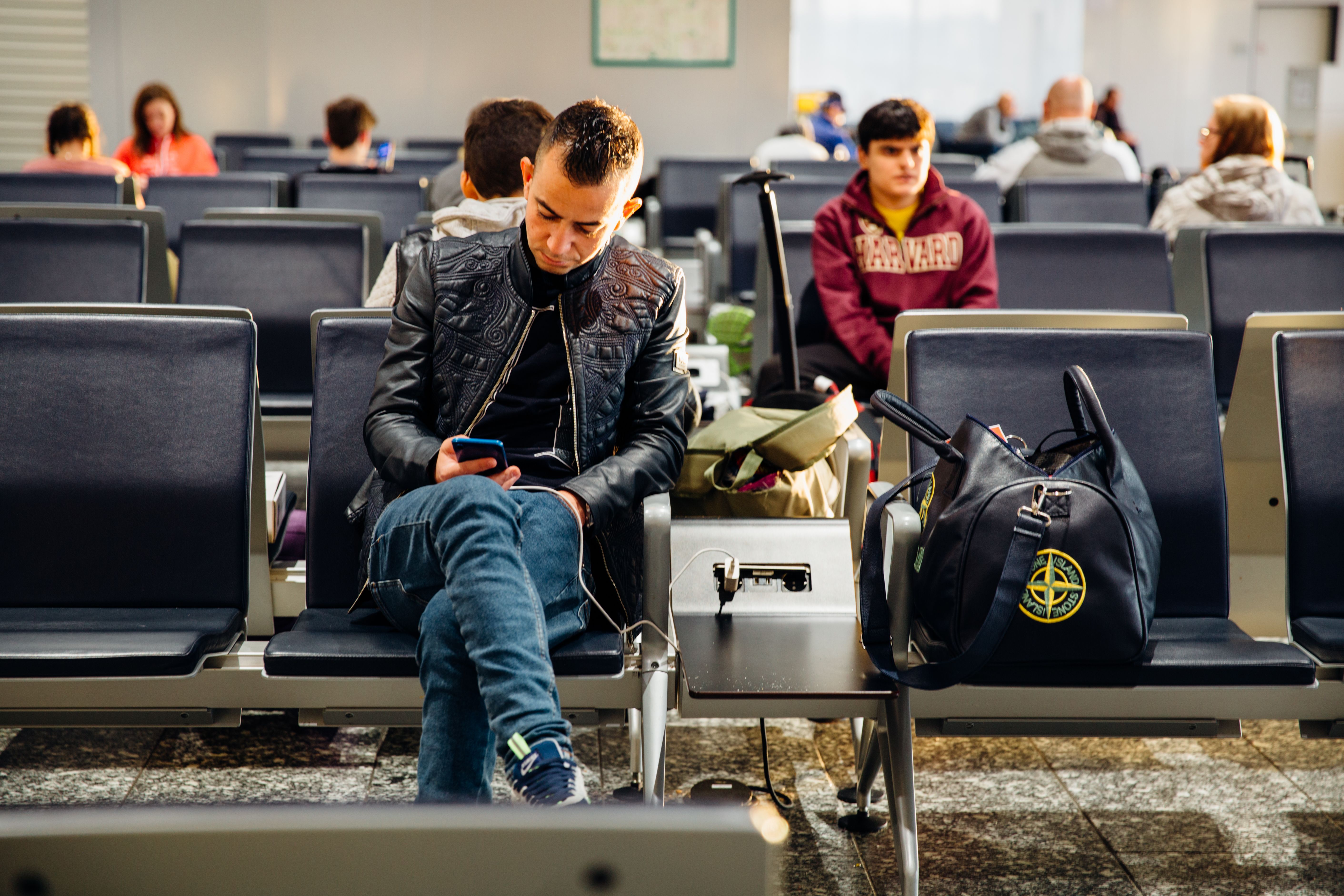 A man sits in an airport gate area and uses his phone while it charges from a port beside his seat. | Source: Pexels