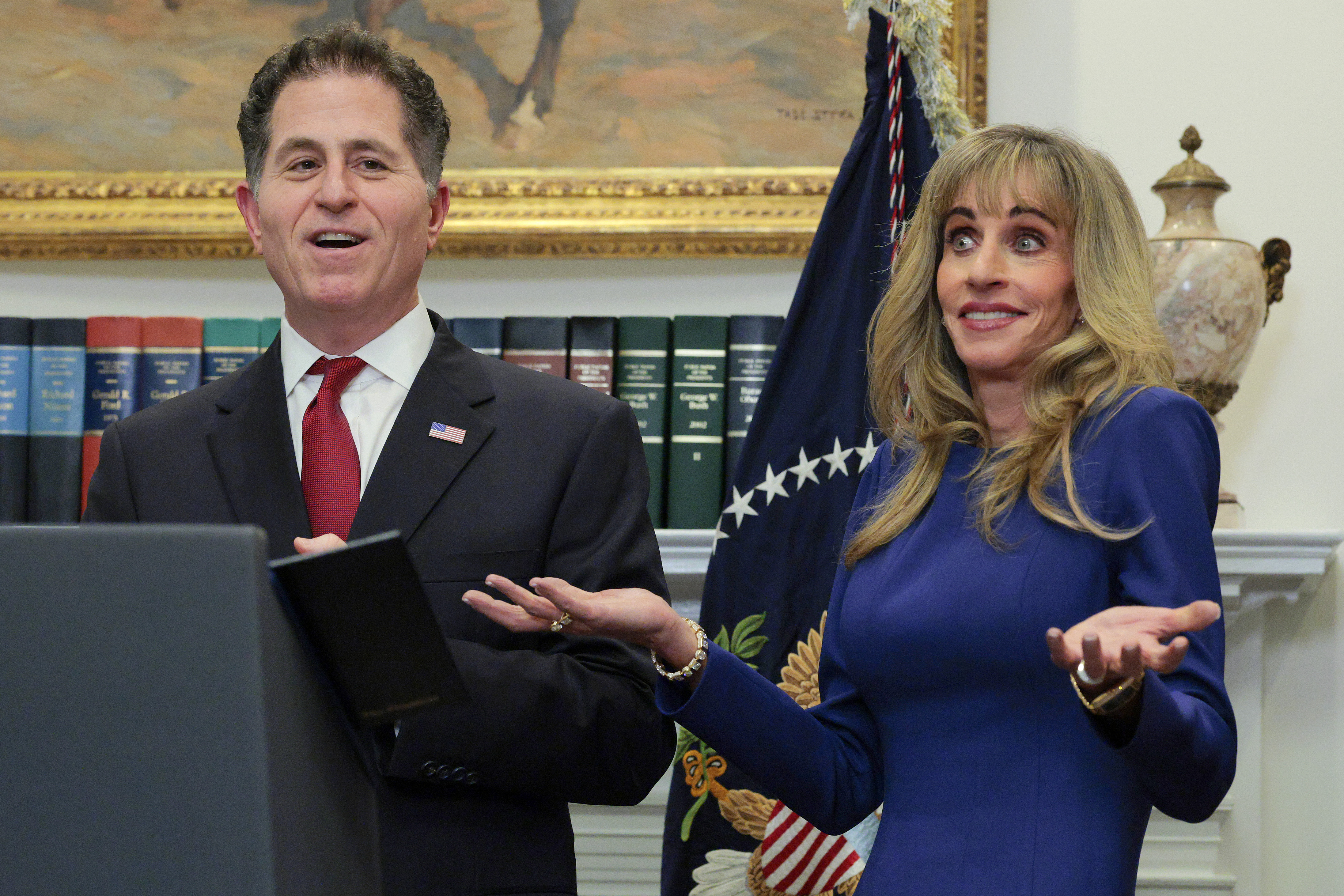 Susan Dell and Michael Dell speak at a lectern in the Roosevelt Room at the White House on December 2, 2025, in Washington, D.C., announcing a $6.25 billion personal contribution toward the "Trump accounts" youth investment initiative. Susan gestures with both hands wearing a cobalt blue long-sleeved dress, bracelet, and stud earrings, standing beside Michael in a dark suit, red tie, and American flag lapel pin. | Source: Getty Images