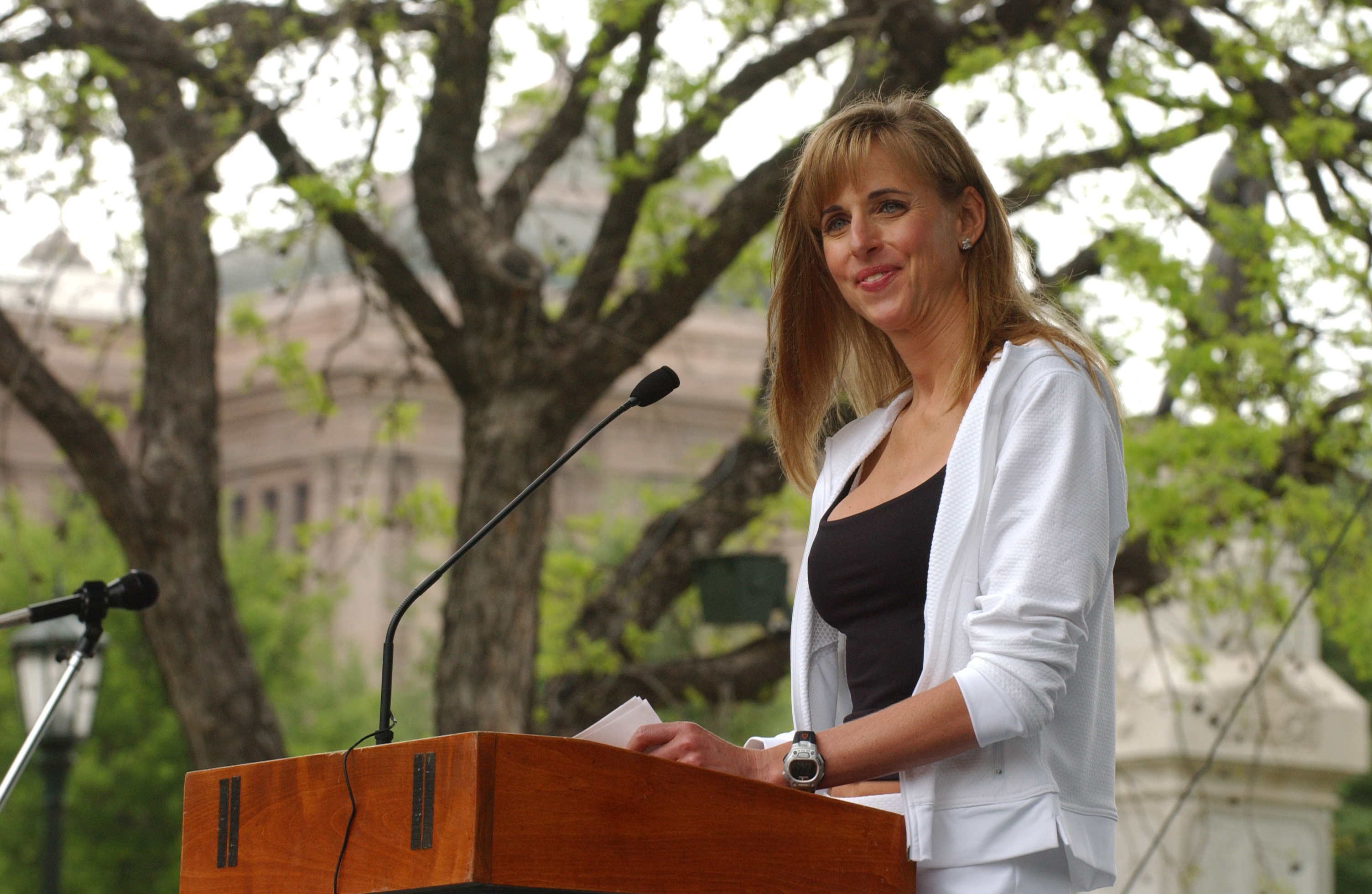 Susan Dell, wife of Dell computers founder Michael Dell and a noted triathlete, addresses the audience from a wooden podium at a fitness fair in Austin on April 17, 2004. She smiles warmly while speaking into the microphone, wearing a sporty white zip-up jacket over a black scoop-neck top, paired with a digital sport watch on her wrist and lush trees visible in the background. | Source: Getty Images