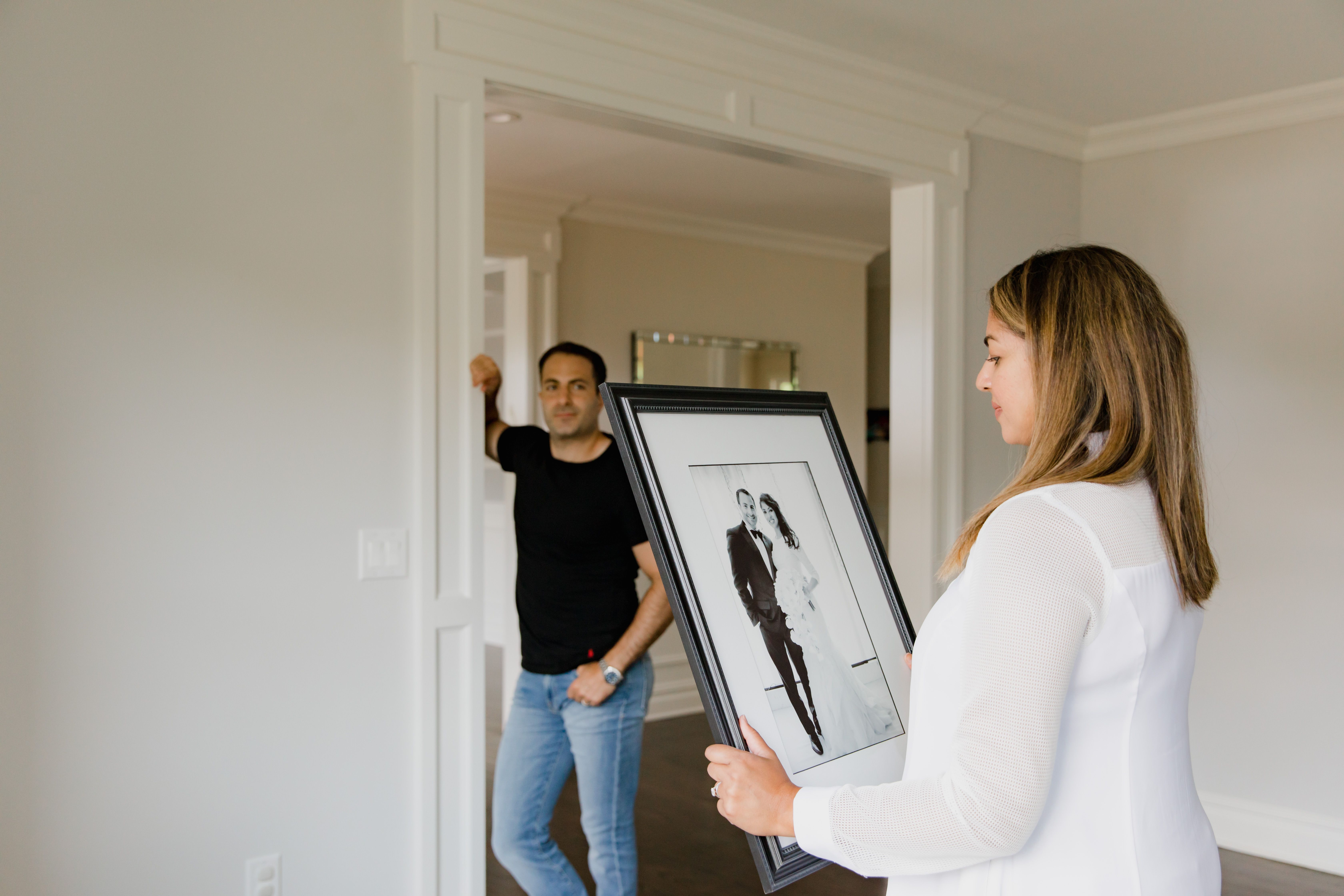 A woman carrying a framed wedding photo while a man watches her while standing by an empty wall | Source: Pexels