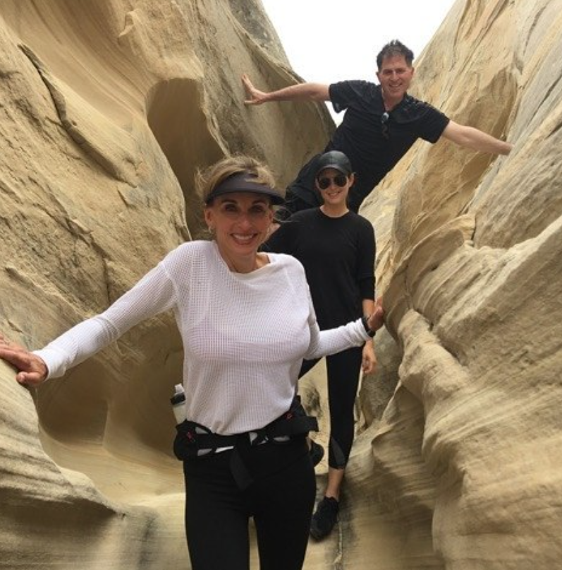Susan Dell, daughter Alexa Dell, and Michael Dell smile while navigating a narrow sandstone slot canyon during an outdoor hiking adventure. Susan leads the way in a white long-sleeved waffle-knit top, black leggings, a sun visor, and a waist pack with a water bottle, while Alexa stands behind her in all-black activewear, dark sunglasses, and a baseball cap, as Michael braces himself playfully between the rock walls above. | Source: Instagram/michaeldell