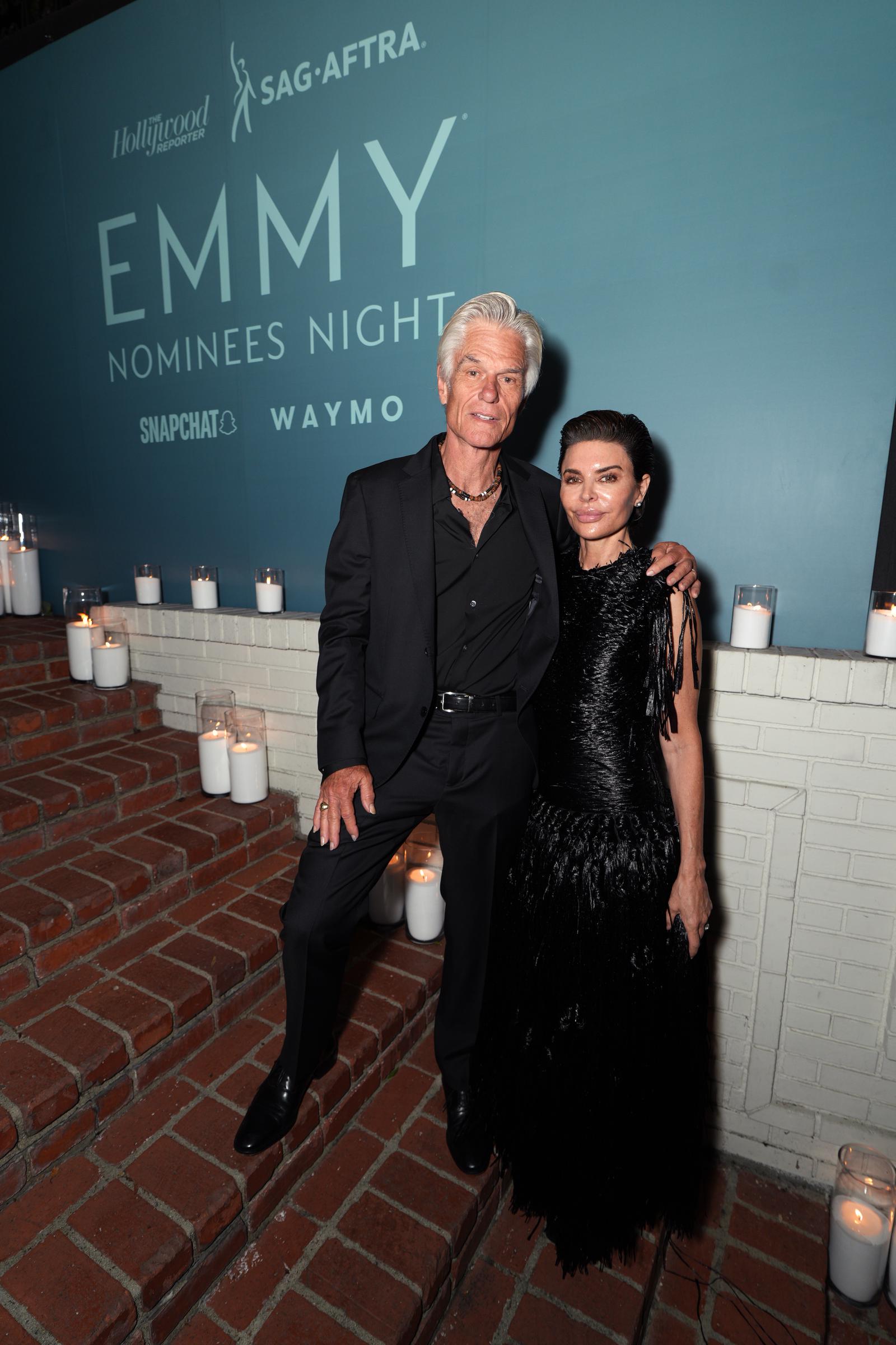 Harry Hamlin puts an arm around Lisa Rinna as they pose together on the candlelit brick steps at The Hollywood Reporter x SAG-AFTRA Emmy Nominees Night in Los Angeles. Rinna’s dramatic black fringe gown creates a striking silhouette beside Hamlin’s all-black look. | Source: Getty Images