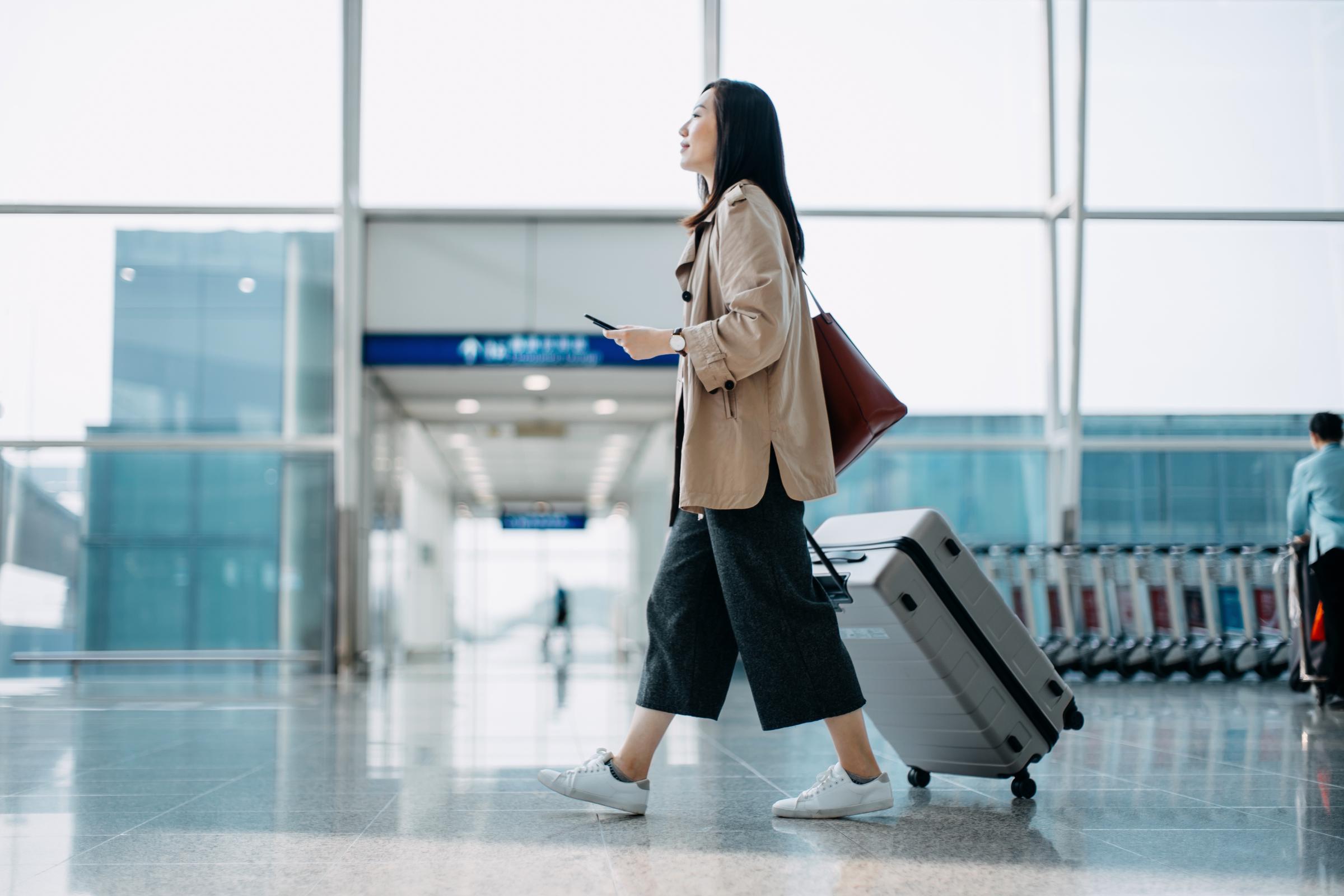 A young woman, carrying a suitcase and holding a smartphone, walks to an airport terminal | Source: Getty Images