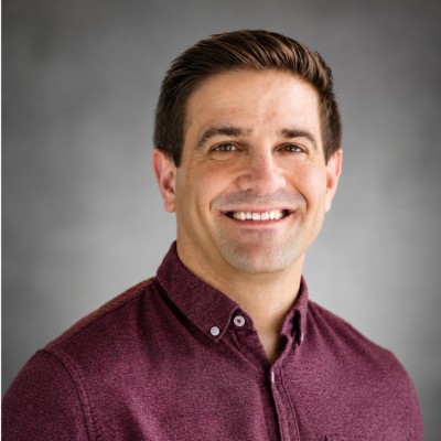 Tyler Zacharia smiles in a headshot portrait set against a neutral grey studio backdrop. He wears a buttoned heather-maroon collared shirt with neat side-parted brown hair. | Source: LinkedIn/Tyler Zacharia