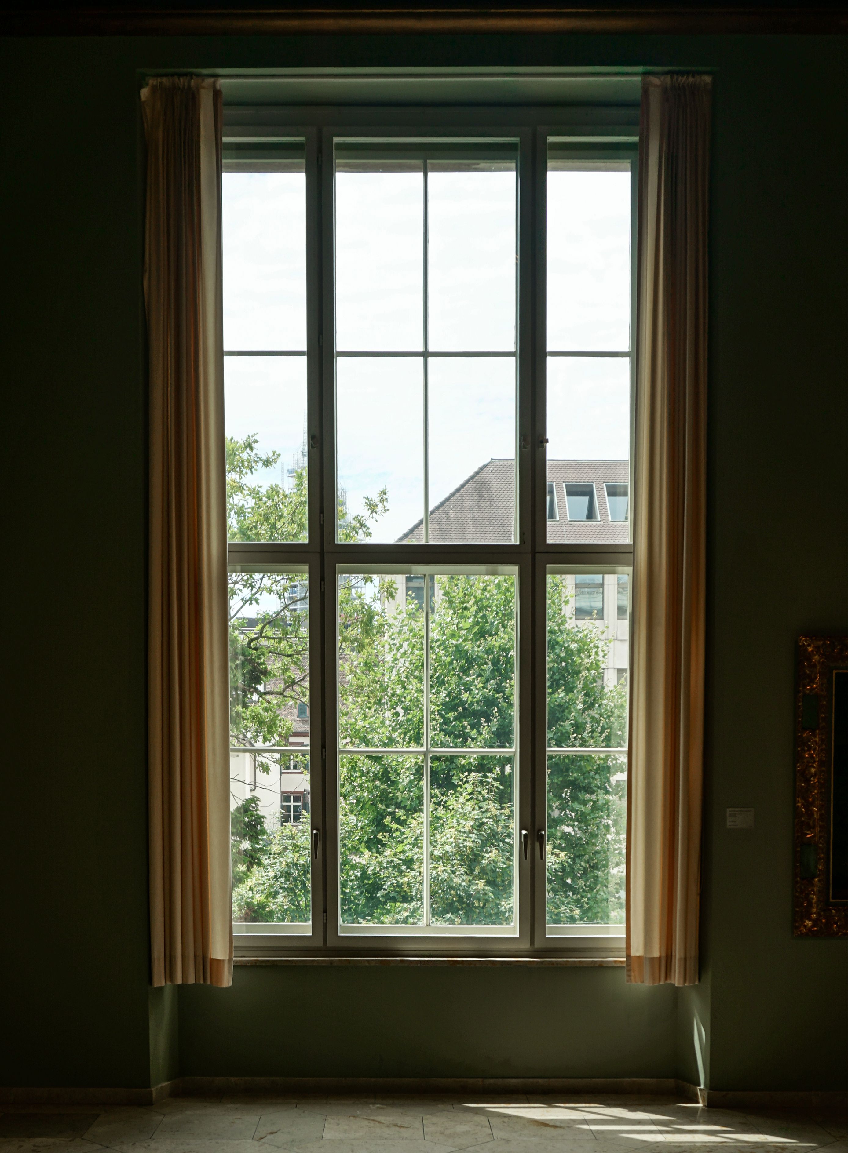 A large window with beige curtains overlooks trees and nearby buildings. | Source: Pexels