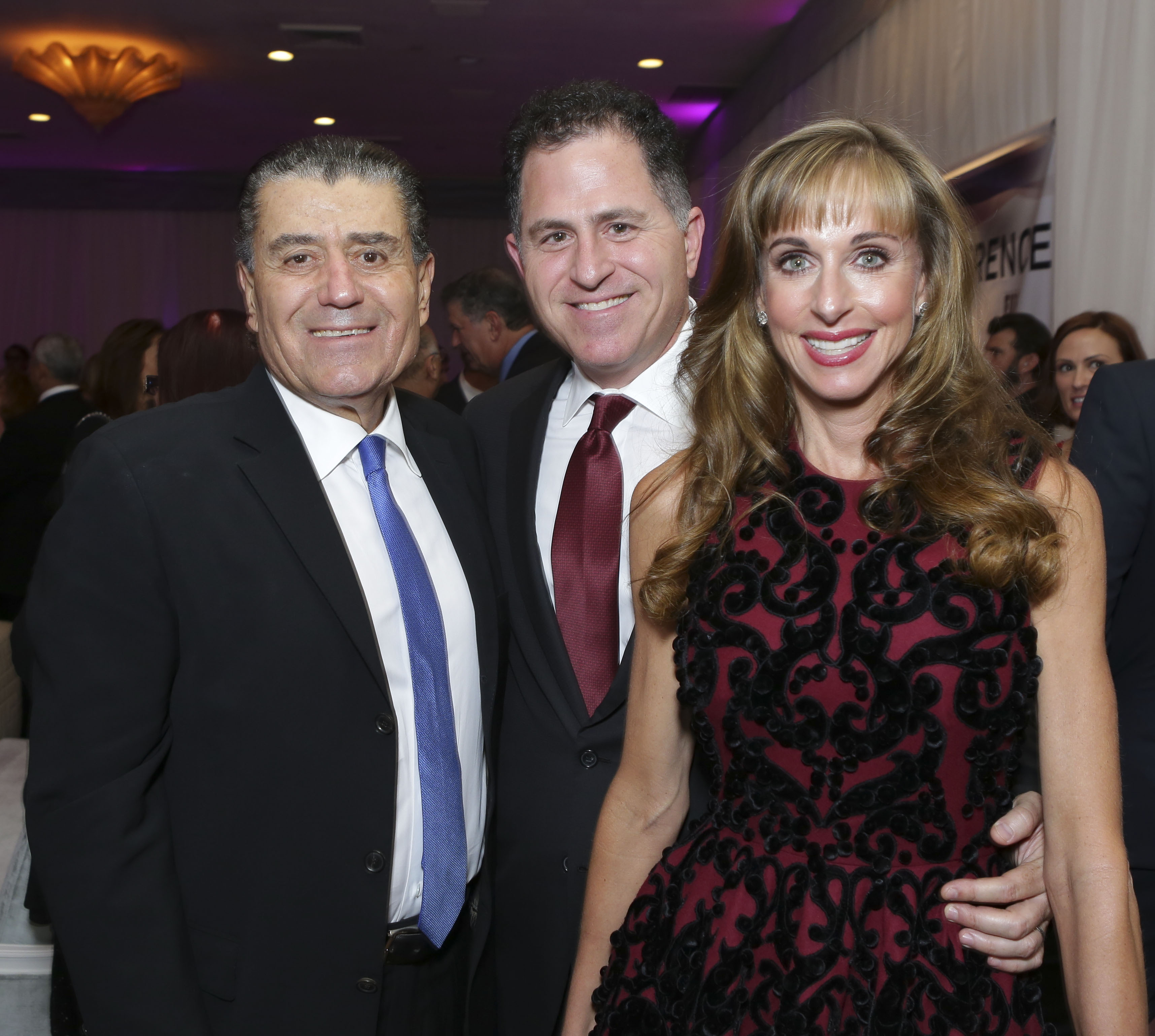 Gala co-chair Haim Saban, Michael Dell, and Susan Dell posed together with bright smiles while attending the Friends of the Israel Defense Forces 2014 Western Region Gala at The Beverly Hilton Hotel on November 6, 2014, in Beverly Hills, California. Saban wore a dark suit with a crisp white shirt and vibrant blue tie, Michael opted for a black suit paired with a deep burgundy necktie, and Susan stood beside them in a sleeveless deep-red dress accented with black velvet scroll embroidery, wearing her hair down in loose curls with neat bangs. | Source: Getty Images