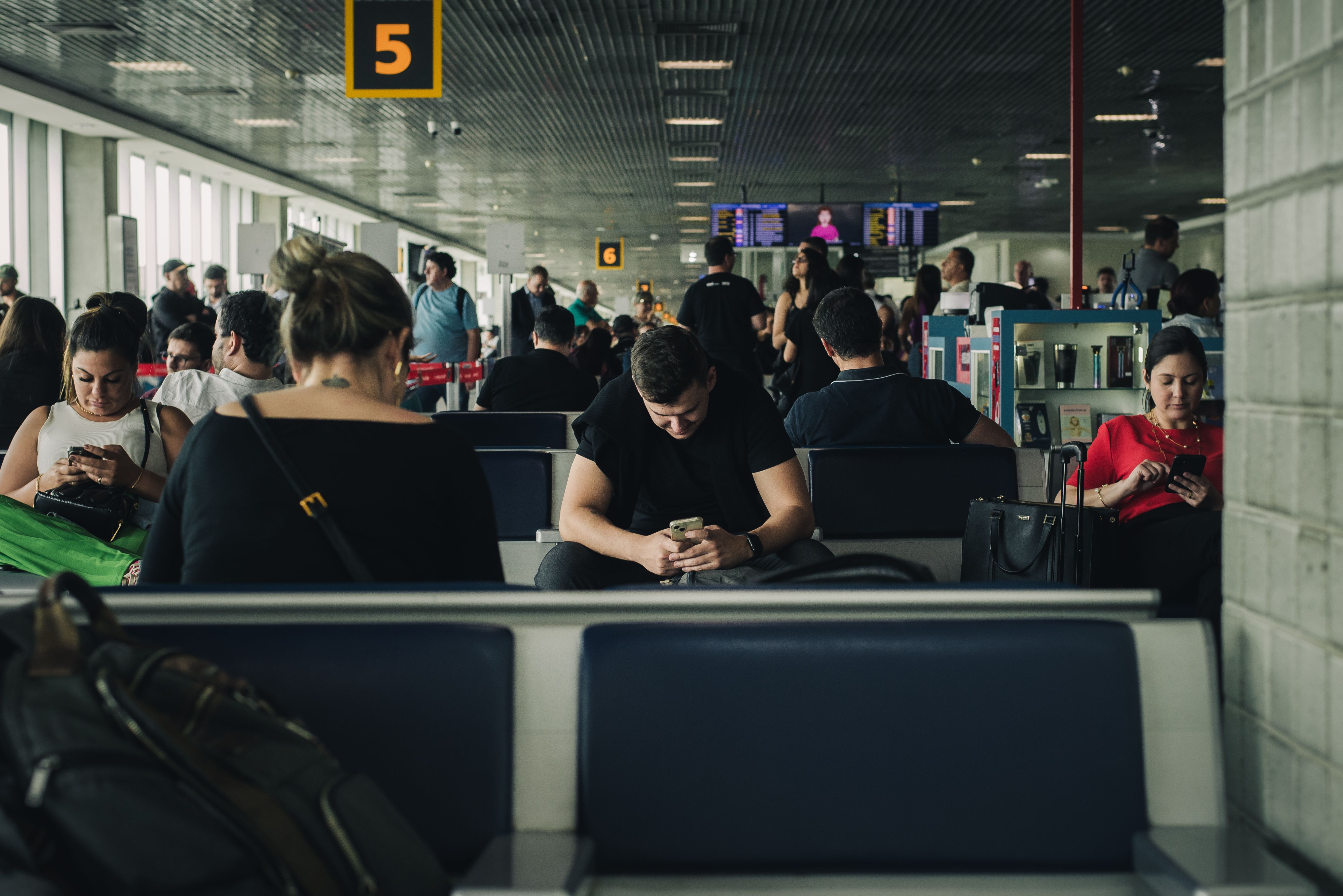 Travelers sit in an airport gate area and look at their phones. | Source: Pexels